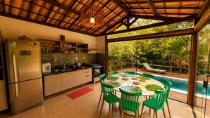 Cucina con frigorifero, piano cottura e lavello, tavolo da pranzo con sedie, e vista su piscina e vegetazione.
