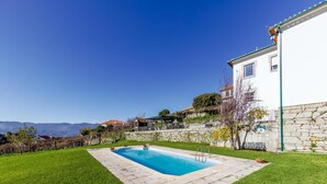 A swimming pool with a stone border, surrounded by a well-maintained lawn, set against a backdrop of a white building and distant hills. - Desassossego House | Swimming pool | Sauna | Peace and quiet (Monção)
