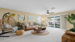 A living room with a sofa, coffee table, and a hanging chair. There are paintings on the wall and a view of the outside through a sliding door. - Your Resort Home! Walk to Disney & Beach! (Kapolei)