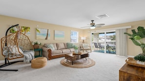 A living room with a sofa, coffee table, and a hanging chair. There are paintings on the wall and a view of the outside through a sliding door. - Your Resort Home! Walk to Disney & Beach! (Kapolei)