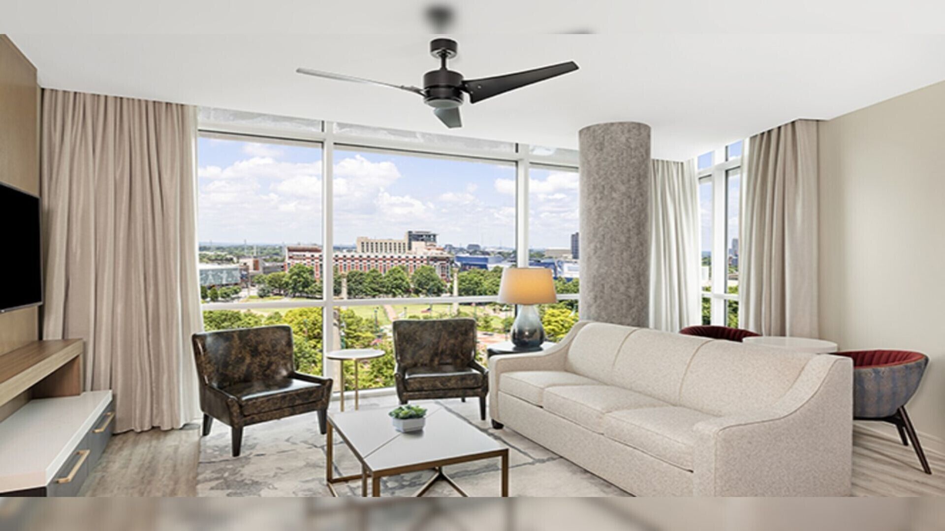 A modern hotel room with a large window, a ceiling fan, a sofa, two armchairs, a coffee table, and a TV.