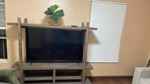 A living room with a wooden entertainment center, a flat-screen TV, a potted plant, and a window with blinds. - Beautiful 3-bedroom townhome with AC in charming Brownsville (Brownsville)