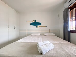 A hotel room with a bed, white curtains, a window with blinds, and a wall-mounted air conditioning unit. - Residencial Mont Sinai (Porto Seguro)