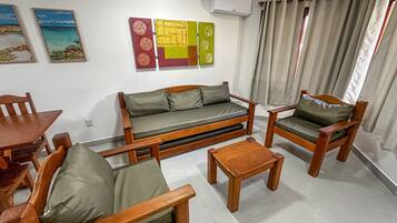 A living room with a sofa, two armchairs, a coffee table, a dining table, and chairs. There are wall-mounted air conditioning units and artwork on the walls.