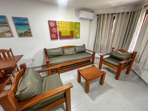 Een woonkamer met een bank, twee fauteuils, een salontafel, een eettafel en stoelen. Er zijn wandgemonteerde airconditioners en kunstwerken aan de muren.