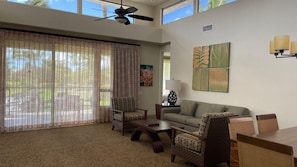 A spacious living room with a large window, a ceiling fan, a sofa, two armchairs, a coffee table, and wall art.