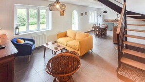 Ein Wohnzimmer mit einer Couch, einem Couchtisch, einem blauen Sessel und einem Korbstuhl. Eine Treppe befindet sich zur Rechten.