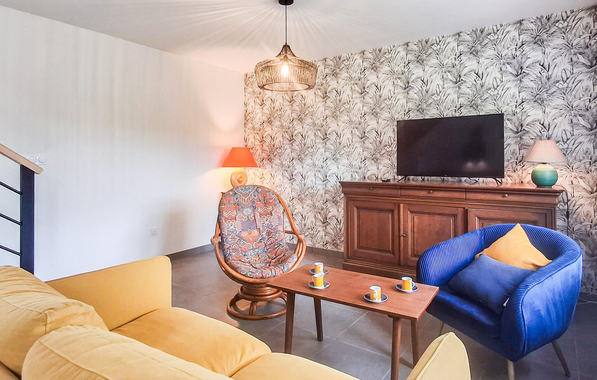 A living room with a sofa, a chair, a table with cups, a TV on a cabinet, and a patterned wall.