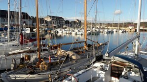 Uma marina com vários veleiros atracados, um edifício com telhado inclinado e água calma refletindo os barcos.