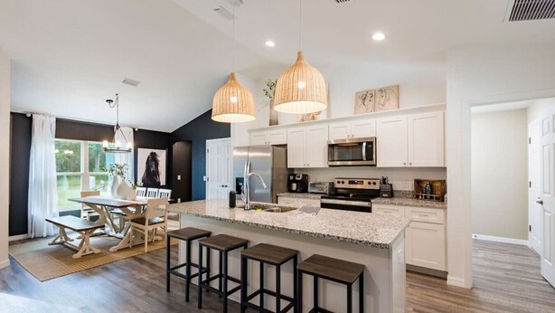 A modern kitchen with white cabinetry, a granite countertop, and stainless steel appliances. There are two hanging pendant lights, a dining area with a table and chairs, and a large island with bar stools. - Springs Basecamp - 4BR Near Rainbow and Manatees (Dunnellon)