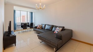 A modern living room with a sectional sofa, a TV on a stand, a dining area with a table and chairs, and a large window with curtains. - Camilo Apartment - By Y Concept (Setúbal)