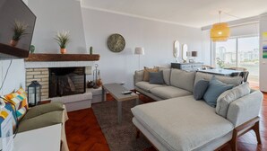 A living room with a fireplace, sofa, and a dining area with a view of the city. - O Ninho dos Sonhos – By Y Concept (Setúbal)