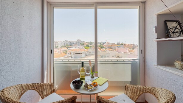 A balcony with wicker chairs, a small table with food and drinks, and a view of the cityscape. - O Ninho dos Sonhos – By Y Concept (Setúbal)
