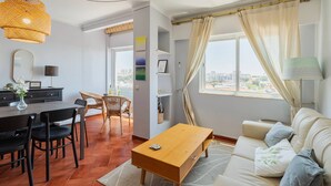 A modern living room with a dining area, a sofa, a wooden coffee table, and a view of the city through large windows. - O Ninho dos Sonhos – By Y Concept (Setúbal)