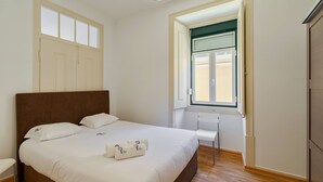 Un lit bien fait, avec du linge de lit blanc et des oreillers, une tête de lit en bois, une petite table de chevet et une fenêtre donnant sur l’extérieur.