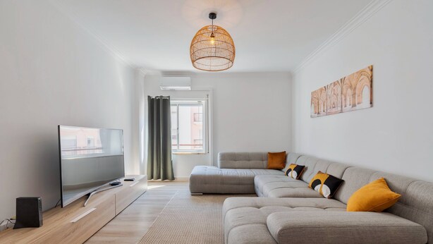 A modern living room with a large flat-screen TV, a grey sectional sofa with mustard pillows, a woven pendant light, and a large window with curtains. - Lucia House - By Y Concept (Setúbal)