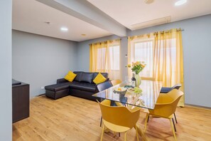 A modern living room with a glass dining table, yellow chairs, a black sofa with yellow cushions, and a large window with yellow curtains.