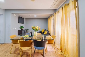 A modern living room with a glass dining table, yellow chairs, a sofa with yellow cushions, and a TV mounted on the wall.