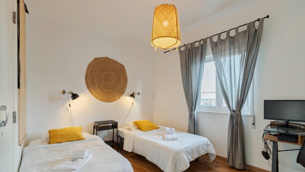 Ett hotellrum med två sängar, ett skrivbord med en dator, en stor vävd väggbonad och ett fönster med gardiner.