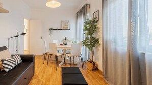 Ett modernt vardagsrum med en svart soffa, ett matbord med stolar, en hängande lampa och ett stort fönster med gardiner.