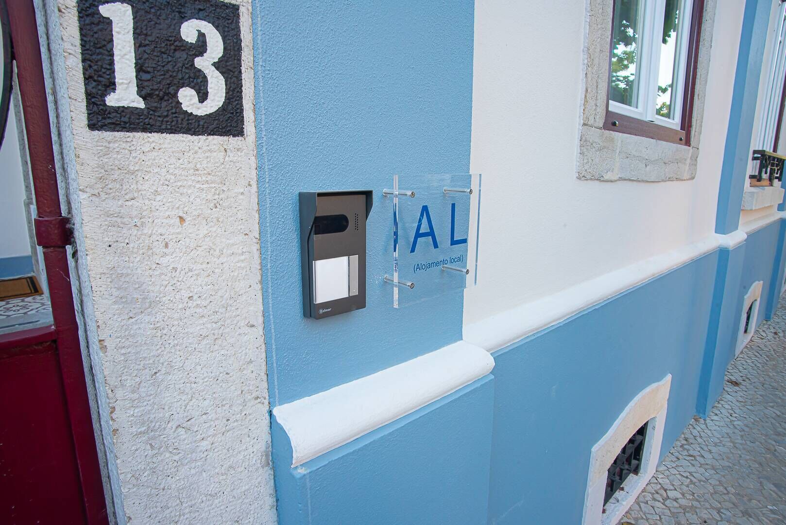 Un bâtiment à la façade bleue et blanche, portant le numéro « 13 » et doté d’une petite fenêtre donnant sur des arbres.