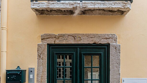 Una puerta verde con base de piedra, un balcón de metal con barandillas ornamentadas y un buzón en la pared.