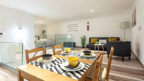 Una sala de estar moderna con un comedor. Hay un mantel a cuadros blanco y negro, sillas de madera y un sofá negro con cojines amarillos. Una pared de cristal separa el comedor de la sala de estar.