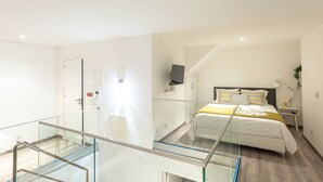 Habitación de hotel moderna con balcón de vidrio, cama con una manta amarilla, televisor montado en la pared y mesita de noche con una lámpara.