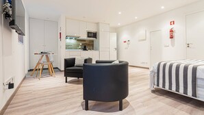 Quarto de hotel moderno com cama, sofá, pequena cozinha e televisão presa à parede.