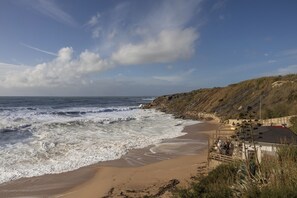 Uma praia com ondas, um penhasco rochoso e um edifício com mesas ao ar livre.