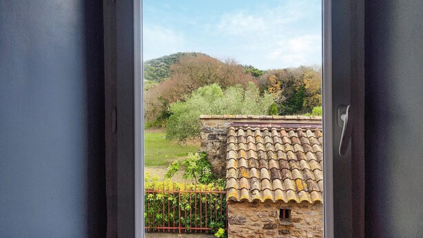 Une vue à travers la fenêtre d’un bâtiment en pierre, doté d’un toit en tuiles, d’une petite fenêtre et d’une clôture en métal rouge au premier plan.