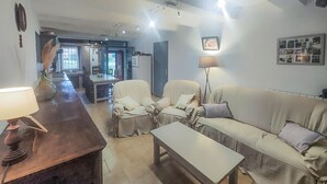 Un salon avec un canapé, des fauteuils, une table basse et une lampe. On aperçoit un coin cuisine au fond.