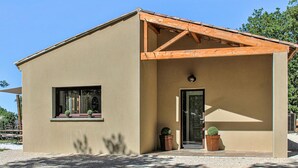 A single-story building with a beige exterior, a wooden gable roof, a small window with potted plants, and a door with a small porch area. - Nice home in Grignan with private swimming pool, can be inside or outside (Grignan)