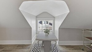 Een kamer met een schuine balkenkap, een raam met zonwering, een tafel met een tafelkleed met patroon, een stoel en een metalen plank.