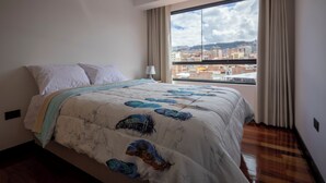 Hotellihuone, jossa on suuri sänky, yöpöytä, lamppu ja suuri ikkuna, josta avautuu näkymä kaupunkiin.