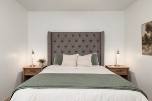 A bedroom with a tufted headboard, a bed with a green blanket, two bedside tables with lamps, and a framed picture on the wall.