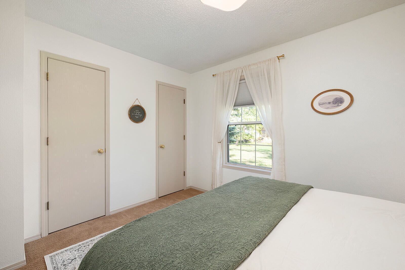 Un dormitorio con cama, ventana con cortinas, puerta y un cuadro colgado en la pared.