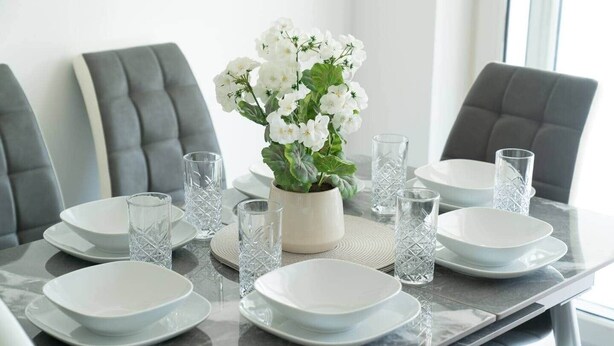 Une table à manger dressée avec de l’argenterie blanche, de la verrerie transparente et une plante en pot ornée de fleurs blanches.