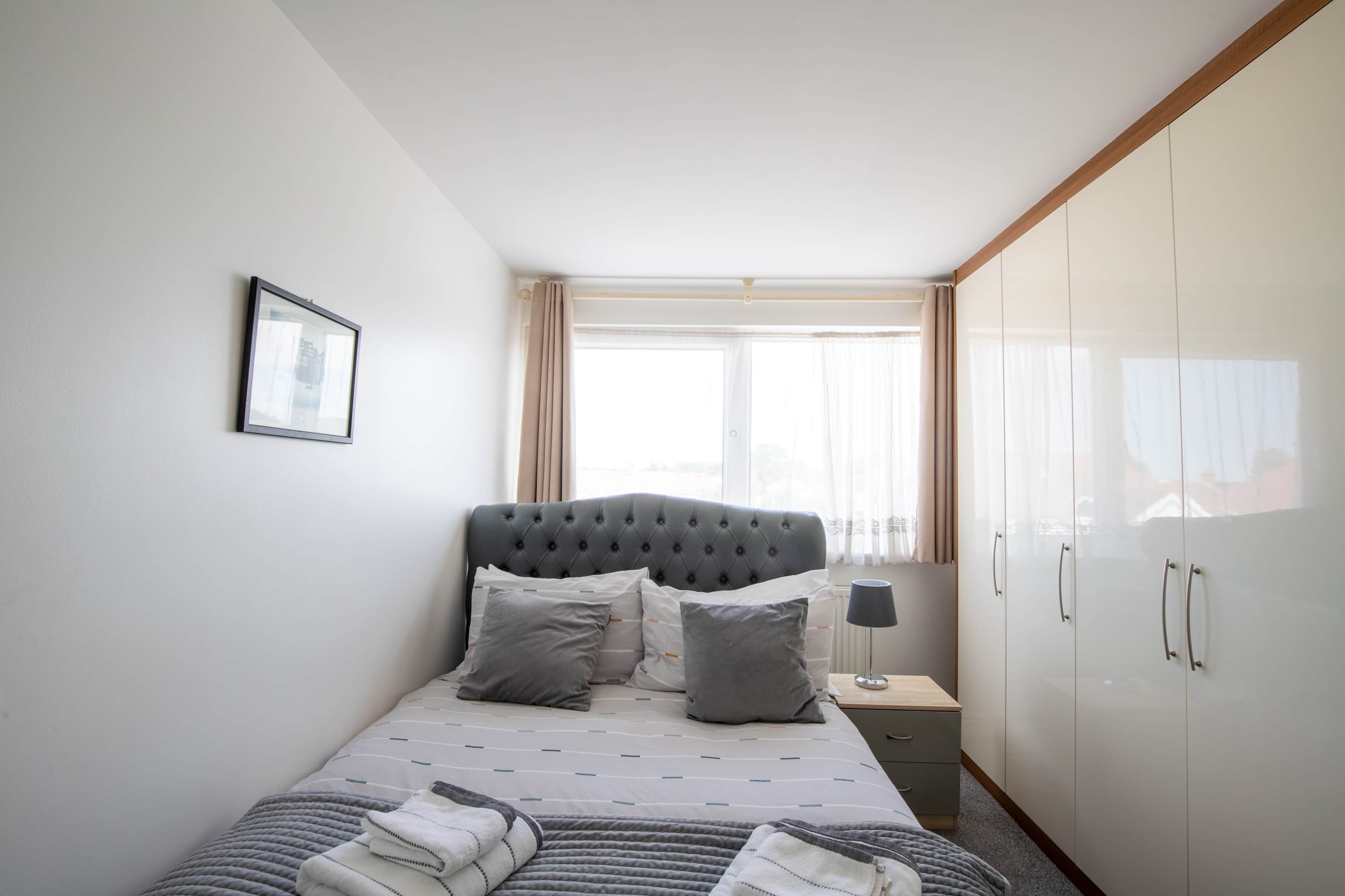 Ein Schlafzimmer mit Bett, Nachttisch, Lampe, Kleiderschrank und einem Fenster mit Vorhängen.
