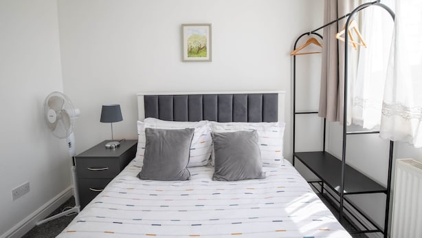 A bedroom with a bed, a bedside table, a lamp, a fan, a radiator, and a window with curtains.