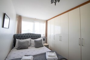 Une chambre à coucher comprenant un lit, une table de chevet, une lampe, une armoire et une fenêtre avec des rideaux.