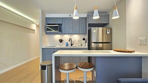 A modern kitchen with grey cabinets, a stainless steel refrigerator, a microwave, and a sink with a herringbone backsplash. There are two stools with wooden seats and white metal frames. - Stunning Studio w/Pool & Includes Free Dedicated Parking Space (Honolulu)