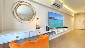 A modern living room with a flat-screen TV mounted on a white console, a round mirror, and a small table with a decorative lamp. - Stunning Studio w/Pool & Includes Free Dedicated Parking Space (Honolulu)
