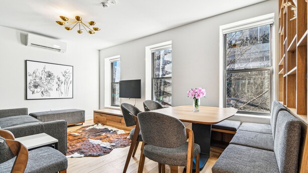 A modern living room with a wooden dining table, grey upholstered chairs, a large window with a view, and a mounted flat-screen TV. - Three Bedroom - Newly Renovated - Washer/Dryer in New York (New York)