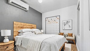 A bedroom with a wooden headboard, a bed with white bedding, a wall-mounted air conditioner, a wall art piece, and a side table with a lamp. - Three Bedroom - Newly Renovated - Washer/Dryer in New York (New York)