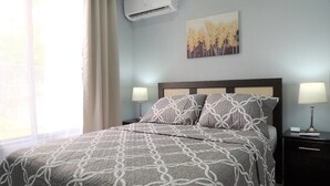 Une chambre à coucher comprenant un lit, deux tables de chevet, un climatiseur fixé au mur et une fenêtre avec des rideaux.