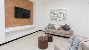 Uma sala de estar moderna com televisão de tela plana fixada em uma parede texturizada, um sofá bege com almofadas e duas mesas laterais redondas com detalhes decorativos.