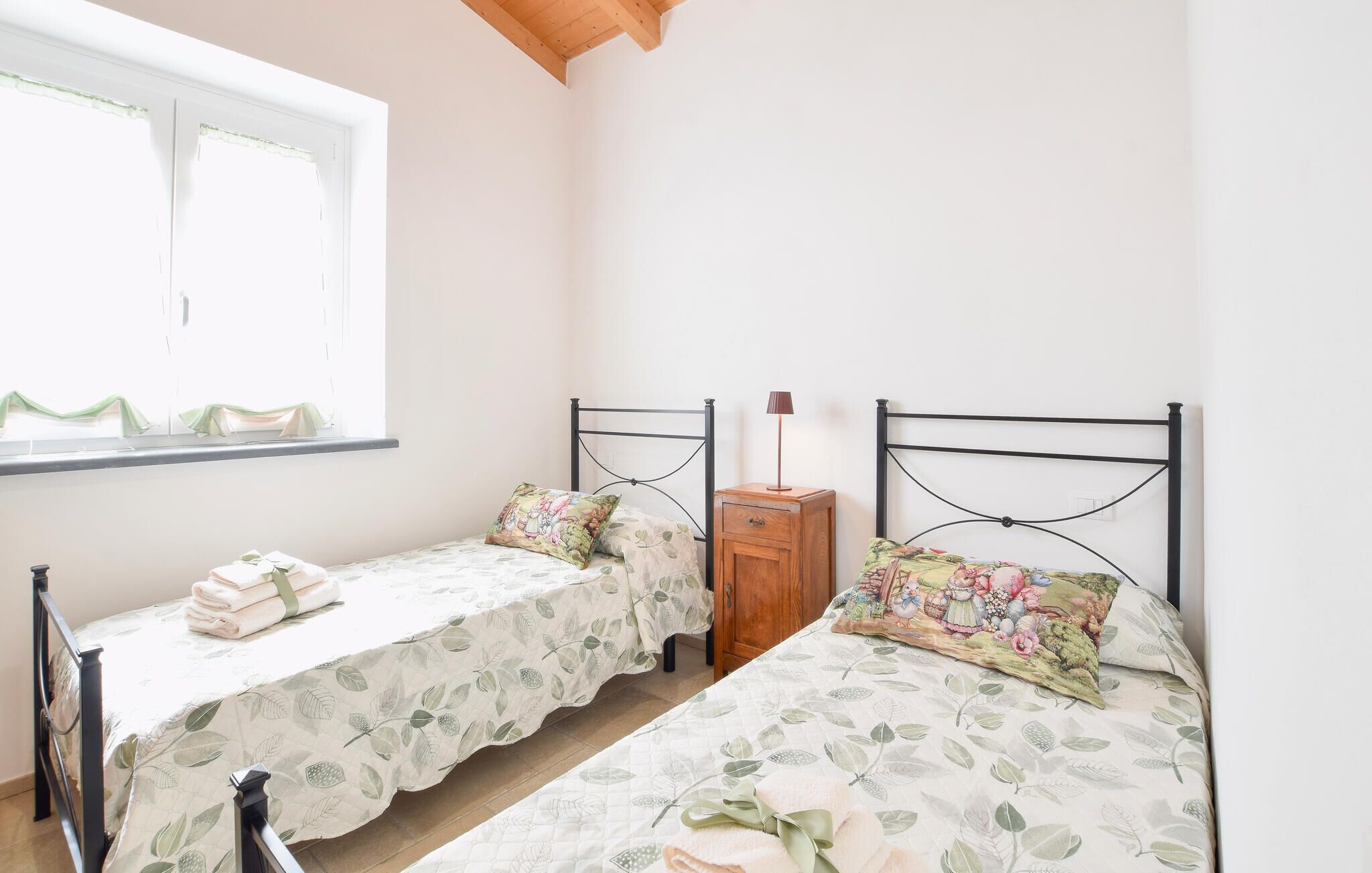 A bedroom with two beds, a window with curtains, a bedside table with a lamp, and a wooden nightstand.