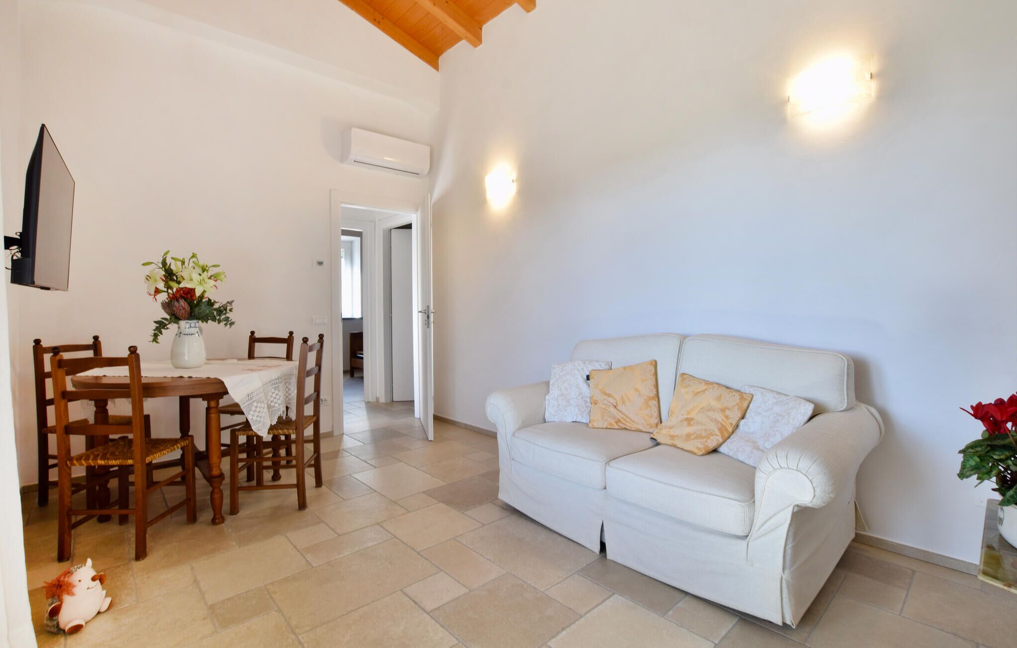 A living room with a white sofa, wooden dining table, and chairs. There is a vase with flowers on the table.