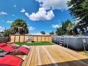 Ein Garten mit einer Holzdachterrasse, einem Grill und einem Pool.